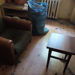 Oddział Czysta Sprawa - Pokój przed sprzątaniem: zniszczony fotel, worek ze śmieciami, stolik i sofa w zabrudzonym pomieszczeniu. Widoczne ślady po zalaniu na podłodze.