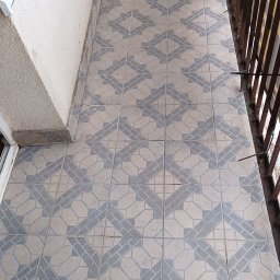 Oddział Czysta Sprawa - Wzór geometryczny na płytkach balkonu, widok z góry. Szare i beżowe płytki tworzą powtarzalny deseń. Fragment ściany i metalowa balustrada po prawej stronie.