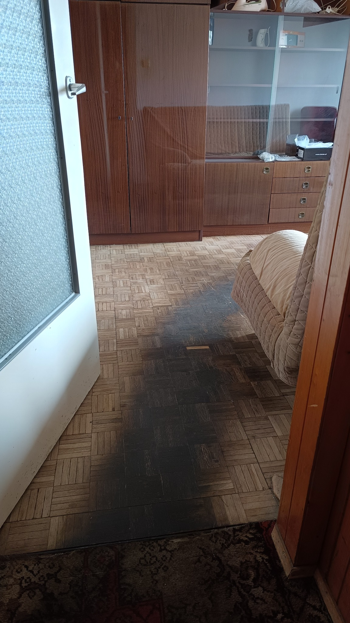 Parkiet po zalaniu, widoczne ciemne plamy. Częściowo widoczny mebel i drzwi. Układ parkietu w kwadraty. Dywan z wzorem w rogu kadru.