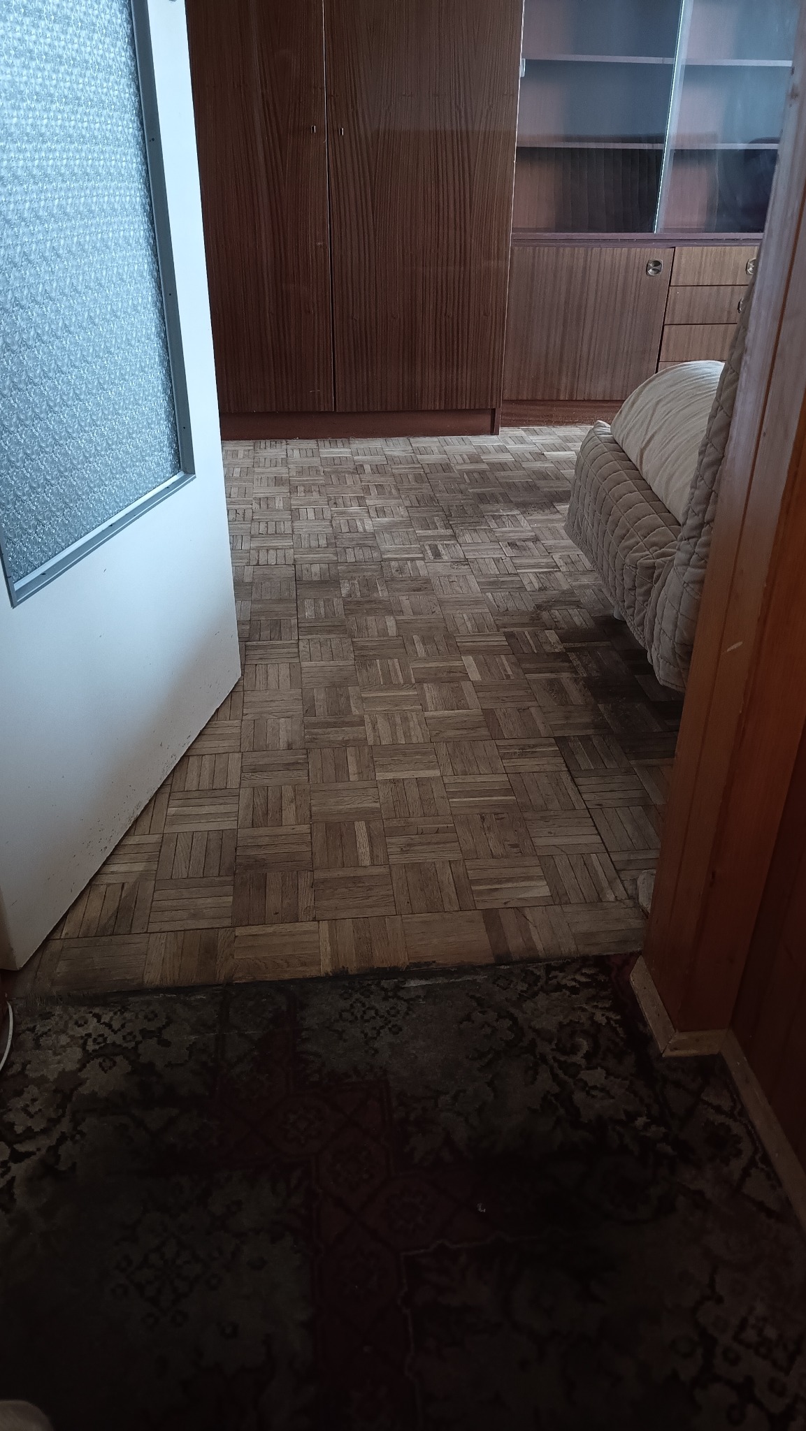 Pokój z parkietem w jodełkę, widok zza otwartych drzwi. W tle szafa i regał z półkami. Dywan w ciemnych barwach w dolnej części kadru.