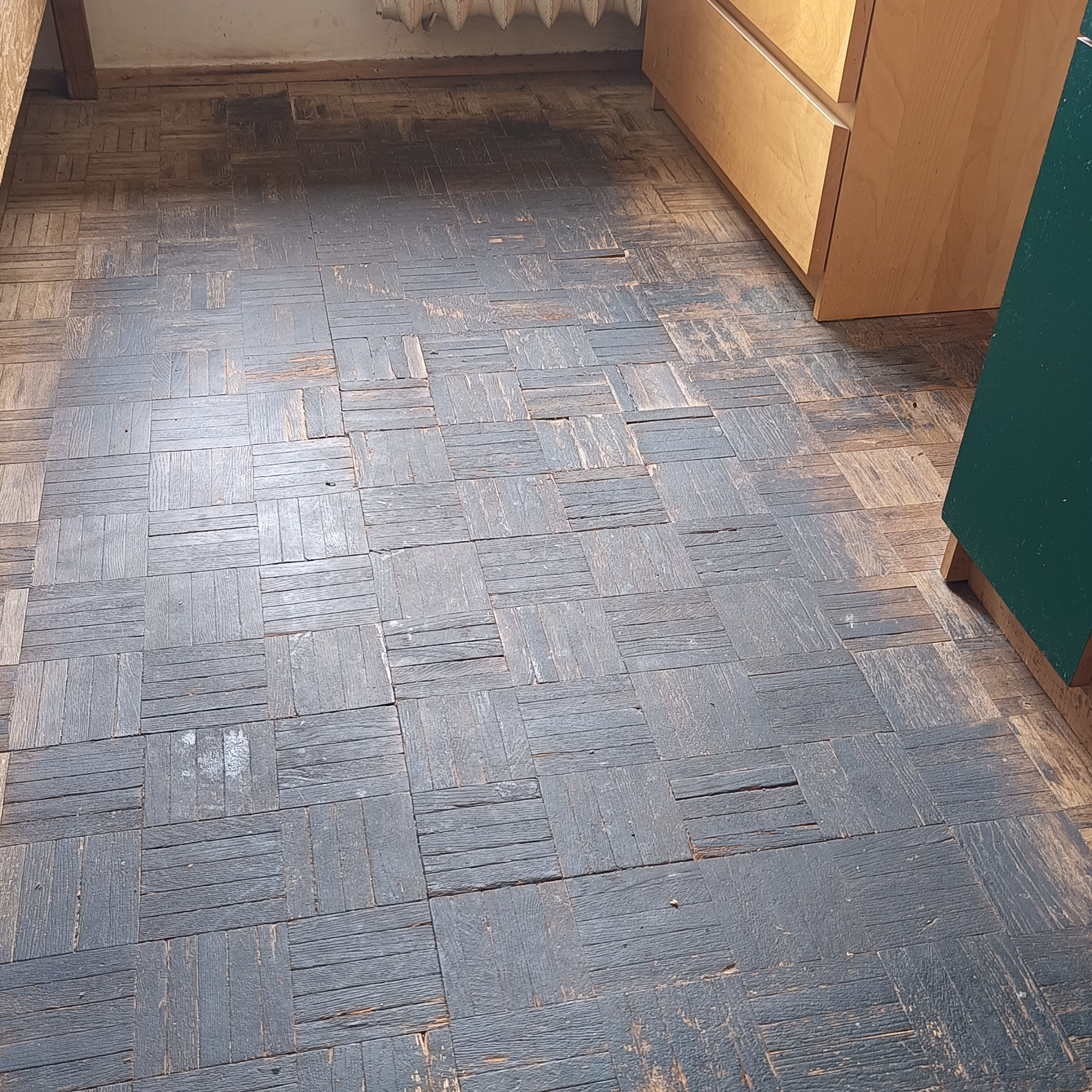 Parkiet z ciemnego drewna ułożony w kwadraty, widoczne ślady użytkowania, rysy i ubytki, przy ścianie szafka z jasnego drewna i fragment zielonej szafki.