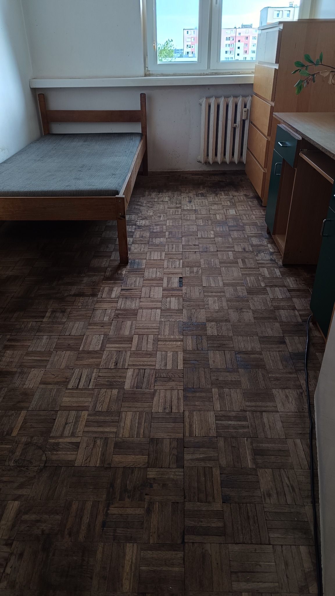 Pokój z parkietem w kwadraty. Widoczne łóżko, grzejnik, szafki i okno z widokiem na budynki. Podłoga wymaga odświeżenia, widoczne zabrudzenia i zarysowania.