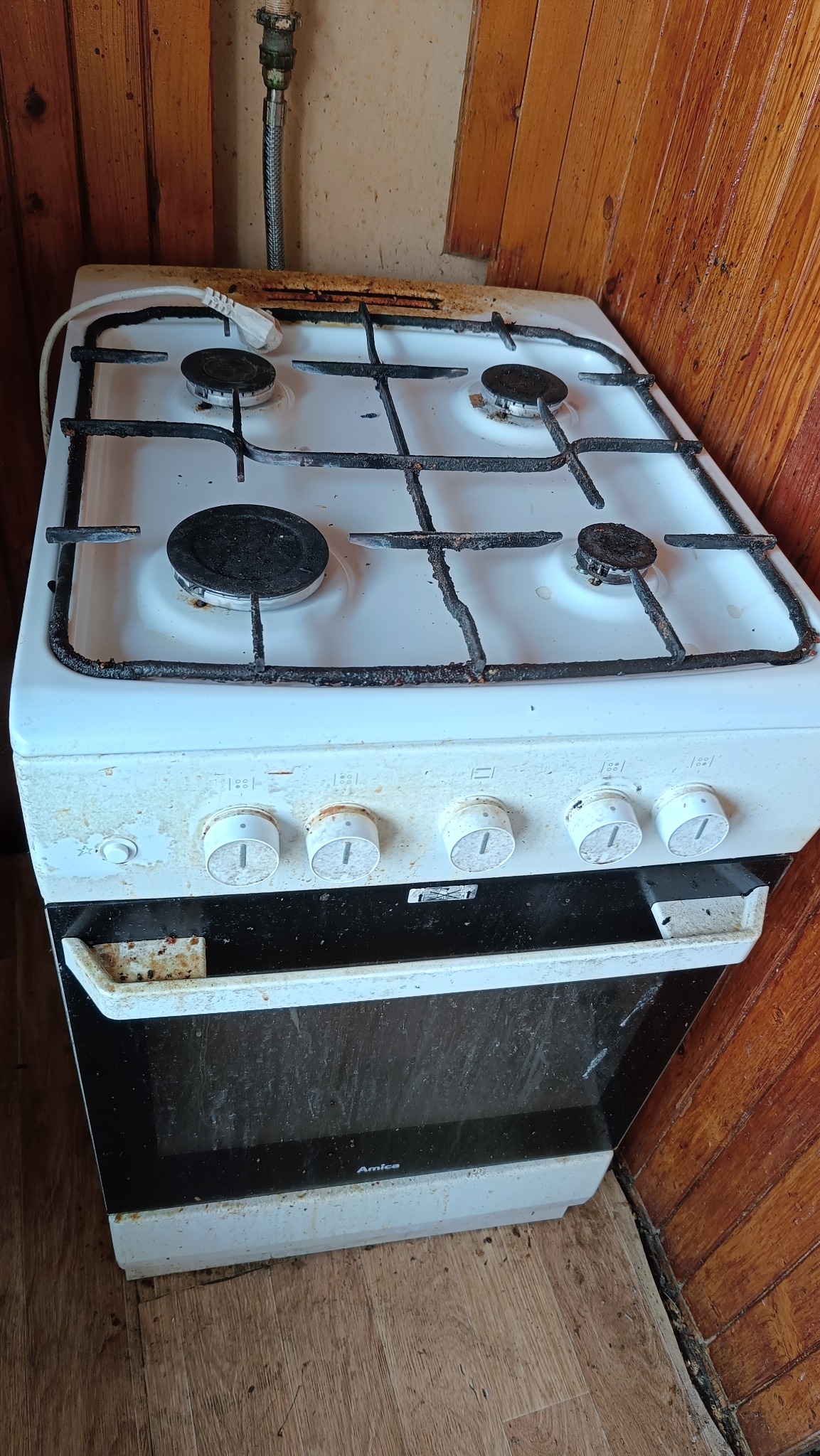 Brudna, biała kuchenka Amica z czterema palnikami na tle boazerii. Widoczne zanieczyszczenia na płycie, ruszcie i pokrętłach. Potencjalne wyzwanie do sprzątania.