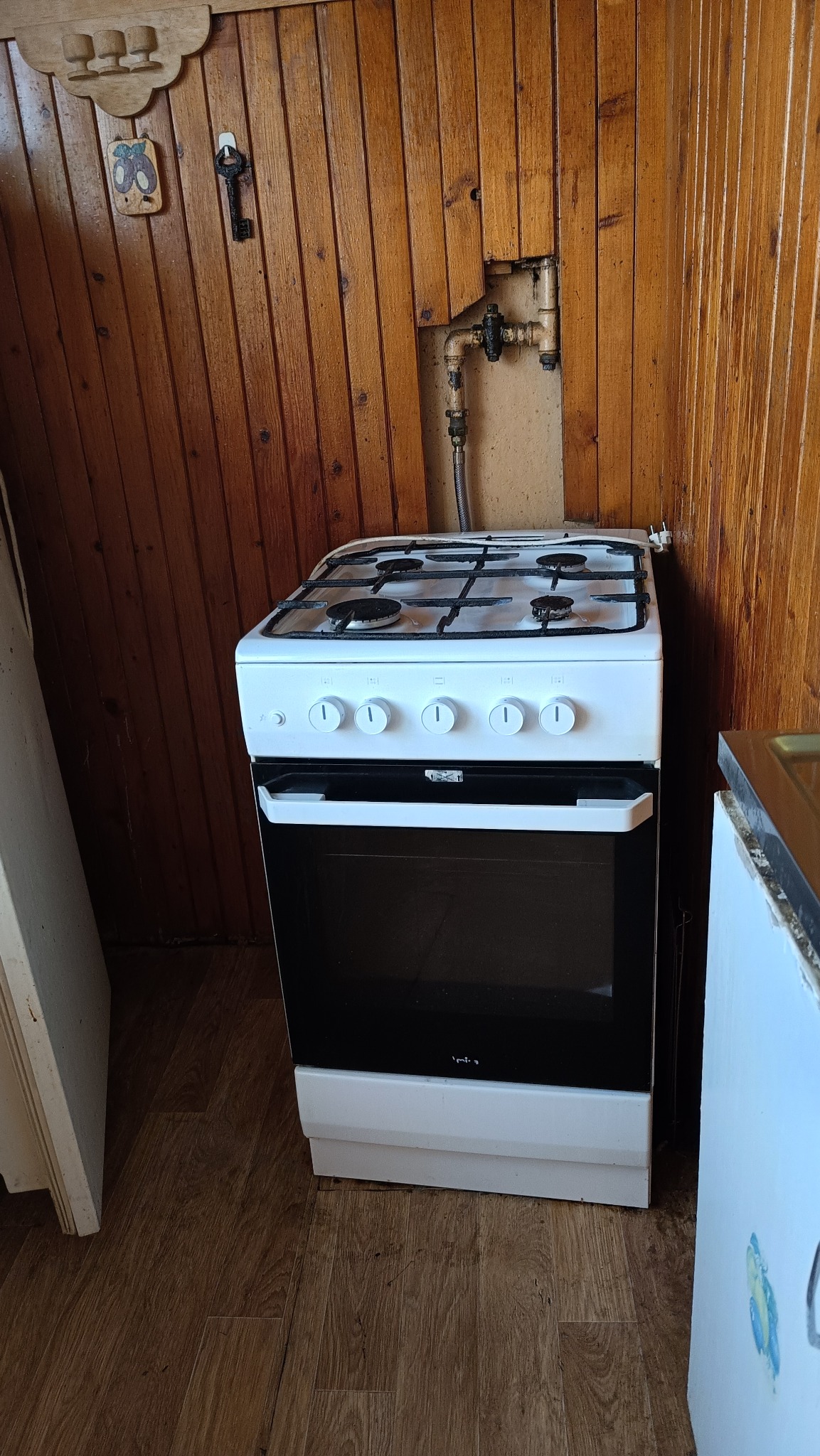 Biała kuchenka gazowa Amica stoi na brązowej podłodze w kuchni wyłożonej boazerią. Nad nią widoczne rury instalacji gazowej i otwór w ścianie.