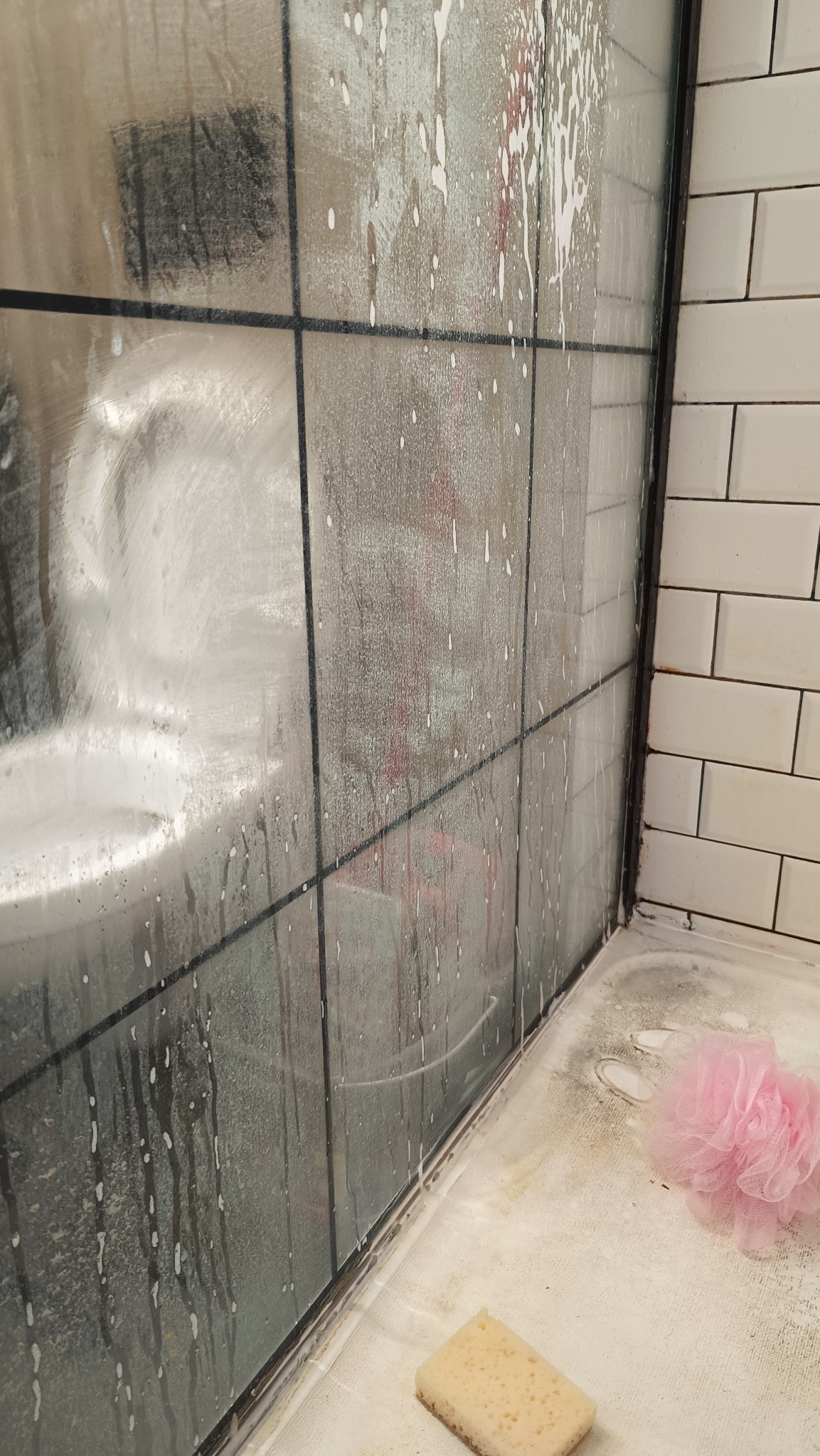 Prysznic z ciemnymi fugami w trakcie czyszczenia. Na szklanej ściance widoczne spływające krople detergentu. Na brodziku leży gąbka i różowa myjka.