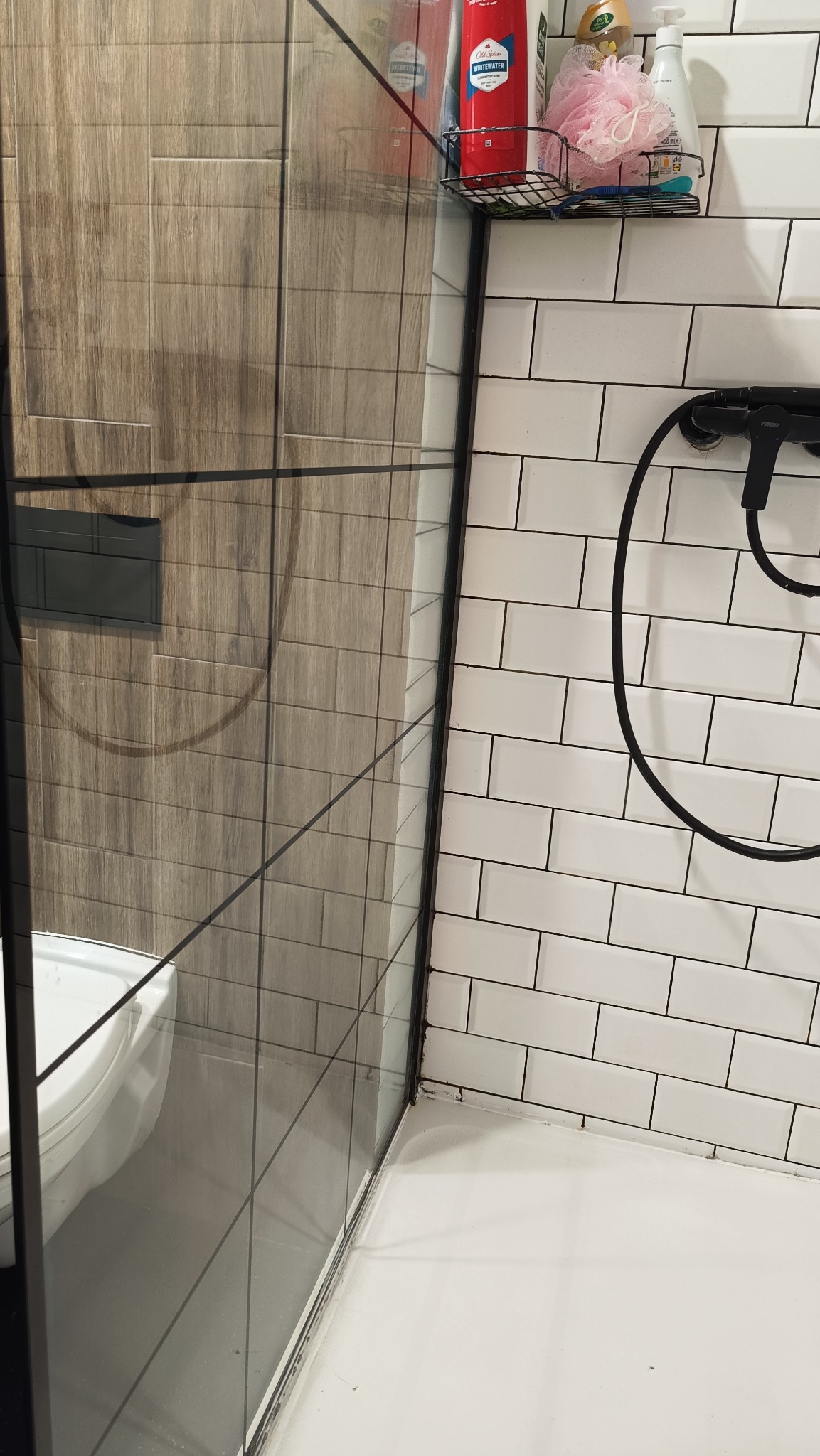 Szczegółowy widok kabiny prysznicowej z czarnymi ramami, białymi płytkami i półką z kosmetykami. Widoczne ślady użytkowania w dolnej części kabiny.