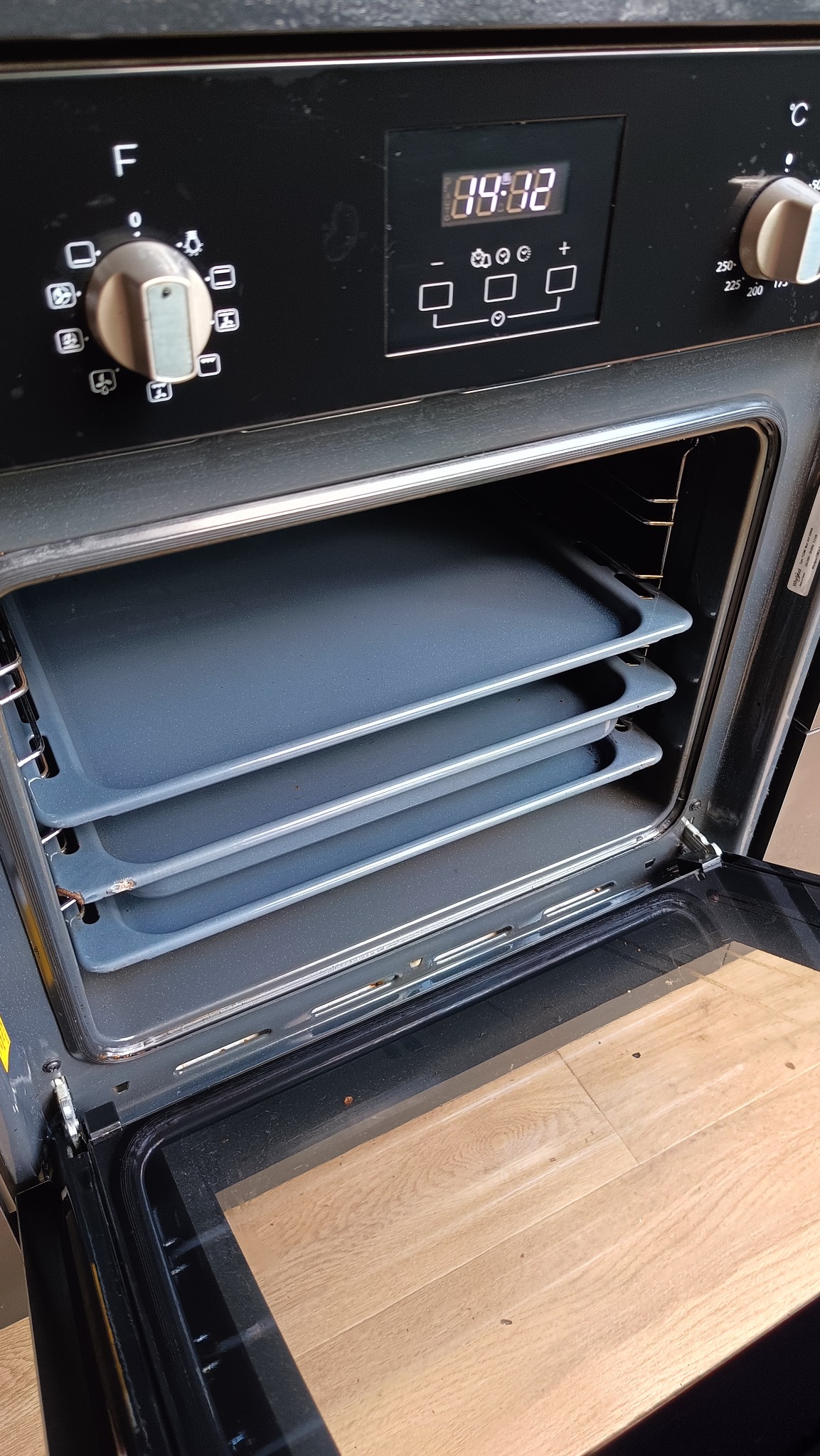 Wnętrze piekarnika z trzema blachami, otwarte drzwiczki, widoczny panel sterowania z wyświetlaczem. Piekarnik w kolorze czarnym, wnętrze szare, podłoga drewniana.