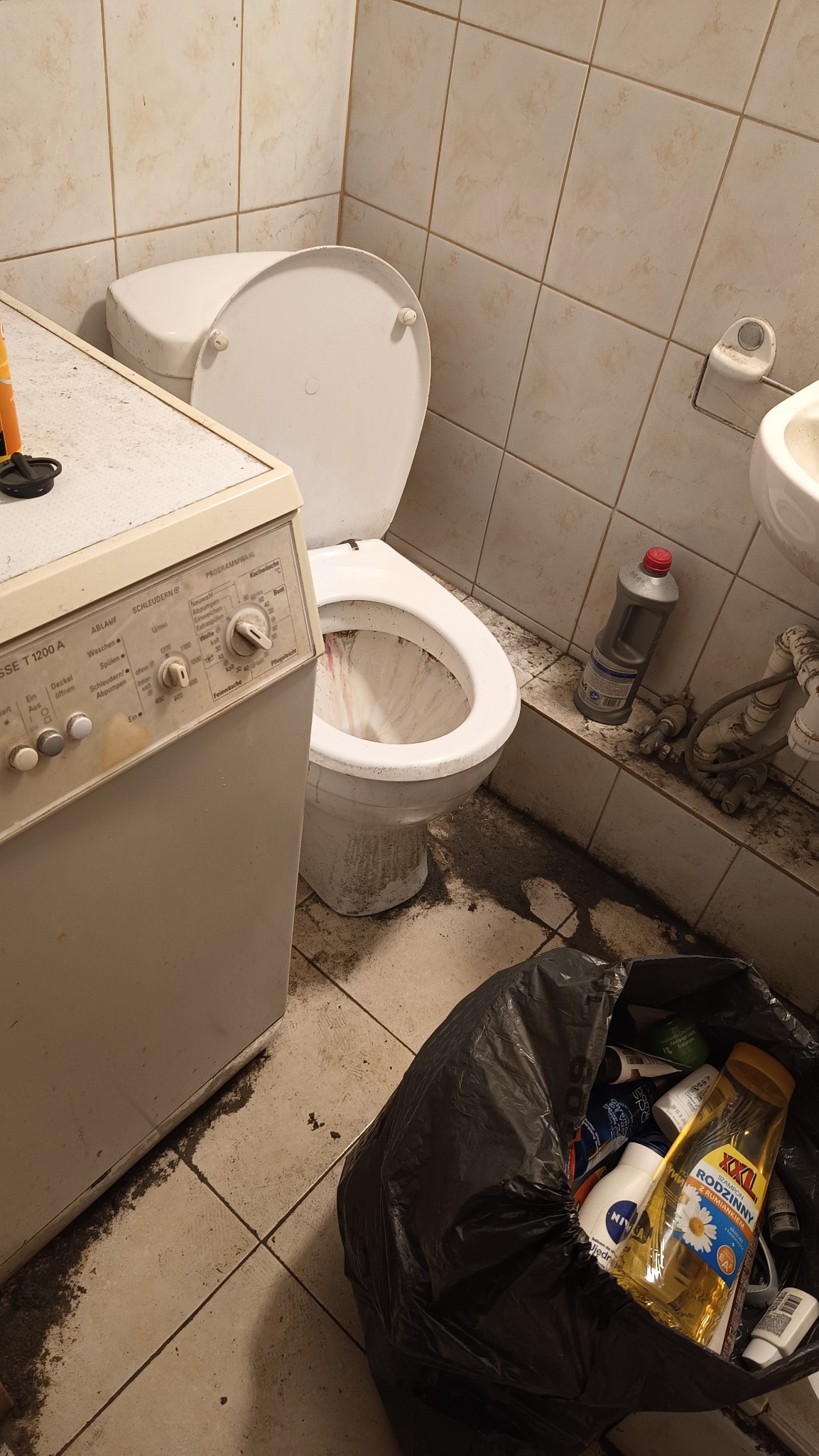 Zaniedbana łazienka z brudną toaletą, starą pralką i workiem na śmieci wypełnionym pustymi opakowaniami po kosmetykach. Widoczne zabrudzenia na podłodze i ścianach.