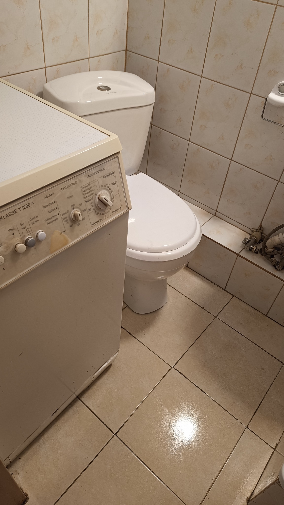 Łazienka z beżowymi płytkami, białą toaletą i starą pralką z widocznymi zabrudzeniami na obudowie i panelu sterowania. Ujęcie z bliska, niskiej jakości.