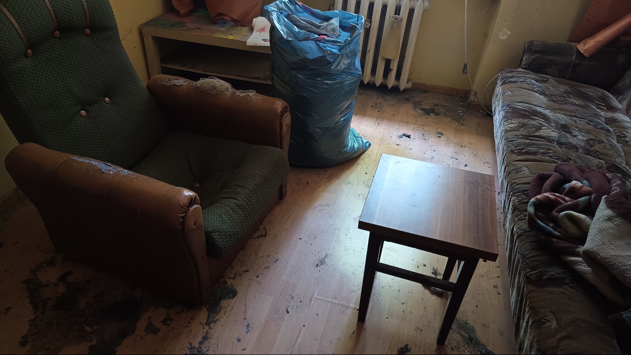 Pokój przed sprzątaniem: zniszczony fotel, worek ze śmieciami, stolik i sofa w zabrudzonym pomieszczeniu. Widoczne ślady po zalaniu na podłodze.