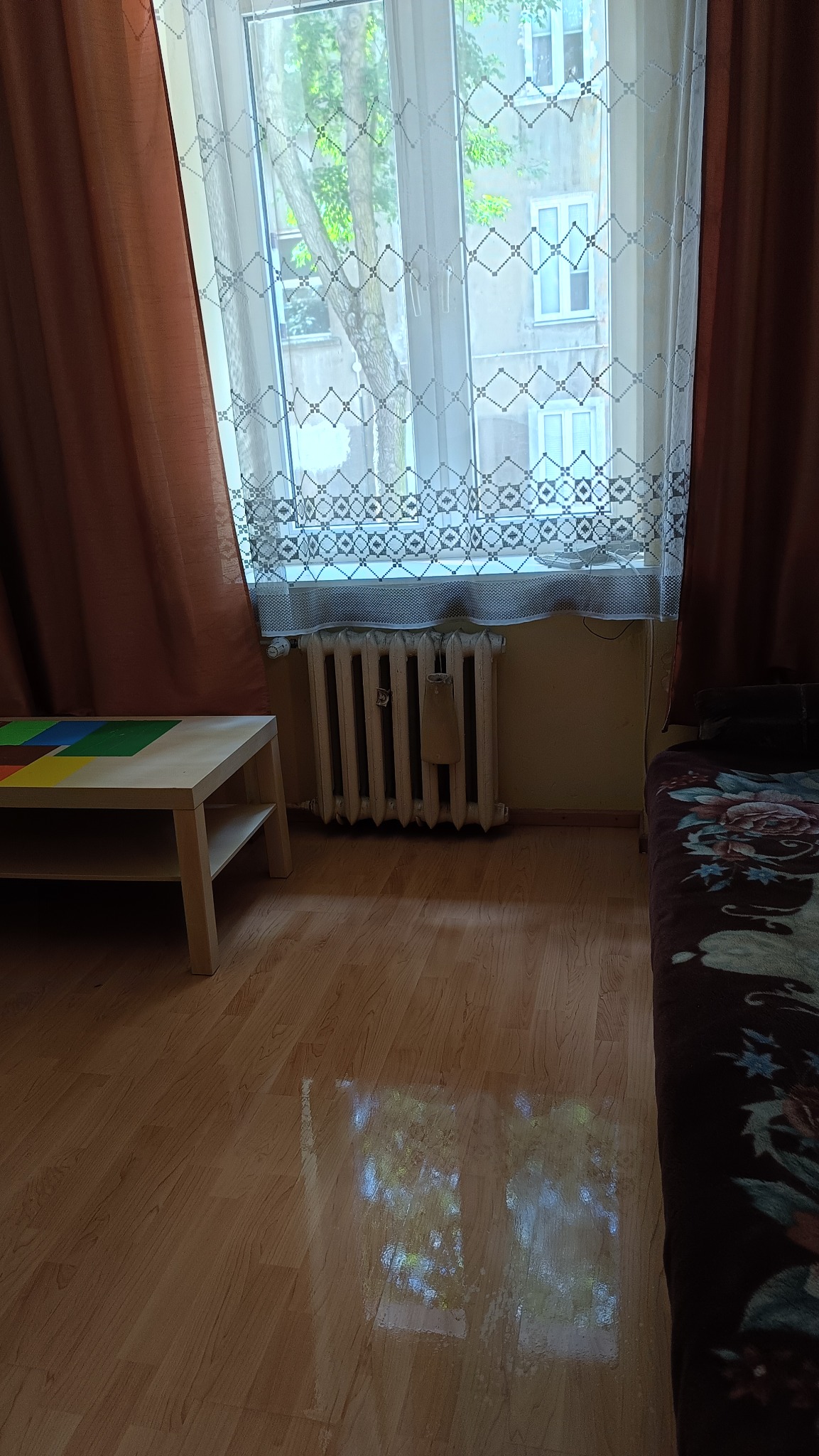 Pokój z widokiem na zieleń za oknem, drewniana podłoga odbija światło. Biały grzejnik pod firanką i stolik z kolorowymi elementami w pokoju.