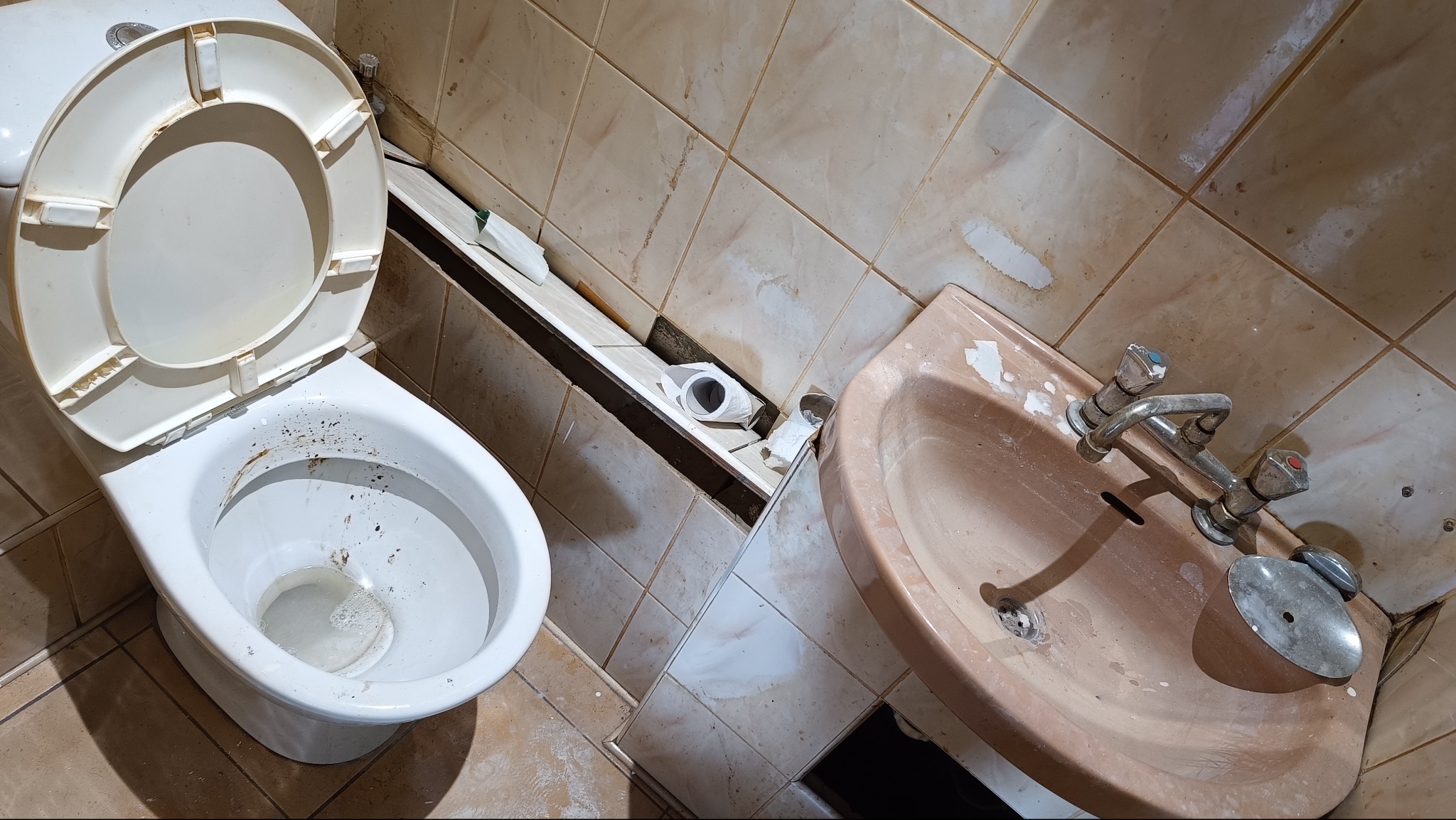 Zaniedbana toaleta z otwartą klapą, brudną muszlą i umywalką. Widoczne uszkodzone płytki i resztki po remoncie. Wnętrze łazienki wymaga gruntownego wysprzątania.