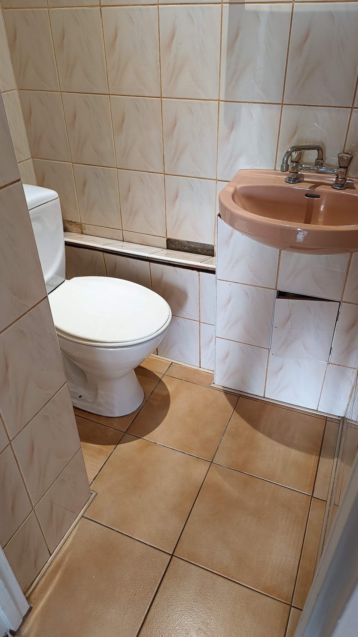 Umywalka z kranem i toaleta w łazience z beżowymi płytkami. Widoczne uszkodzenia na ścianie i podłodze. Zaniedbana łazienka przed sprzątaniem lub remontem.