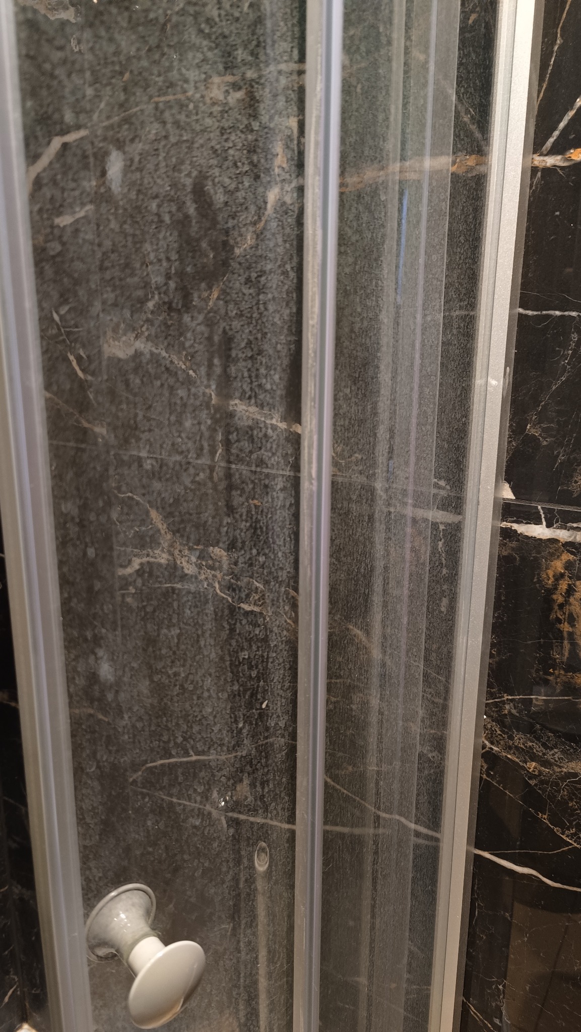 Zabrudzona kabina prysznicowa z osadem z kamienia na szklanych drzwiach i ciemnych płytkach imitujących marmur. Widoczny uchwyt do otwierania drzwi.