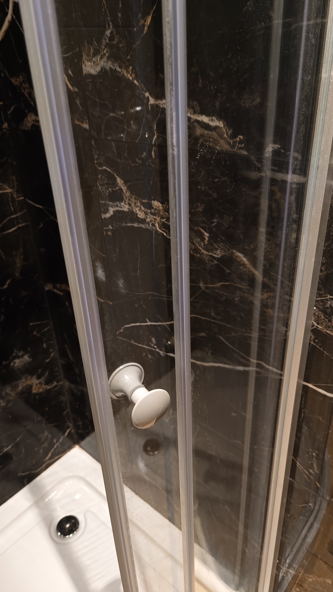 Szczegółowe ujęcie kabiny prysznicowej z widocznymi zaciekami na szkle i czarnymi płytkami imitującymi marmur w tle, uchwyt kabiny w kolorze białym.