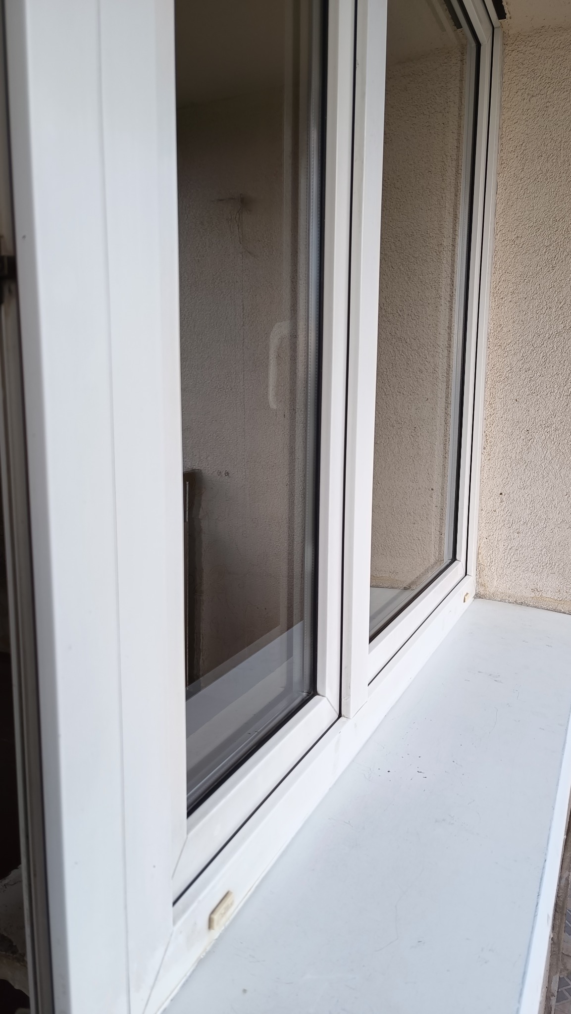 Białe okno PCV z widocznym brudnym parapetem i smugami na szybie, uchwycone pod kątem, ukazujące potrzebę umycia okien.