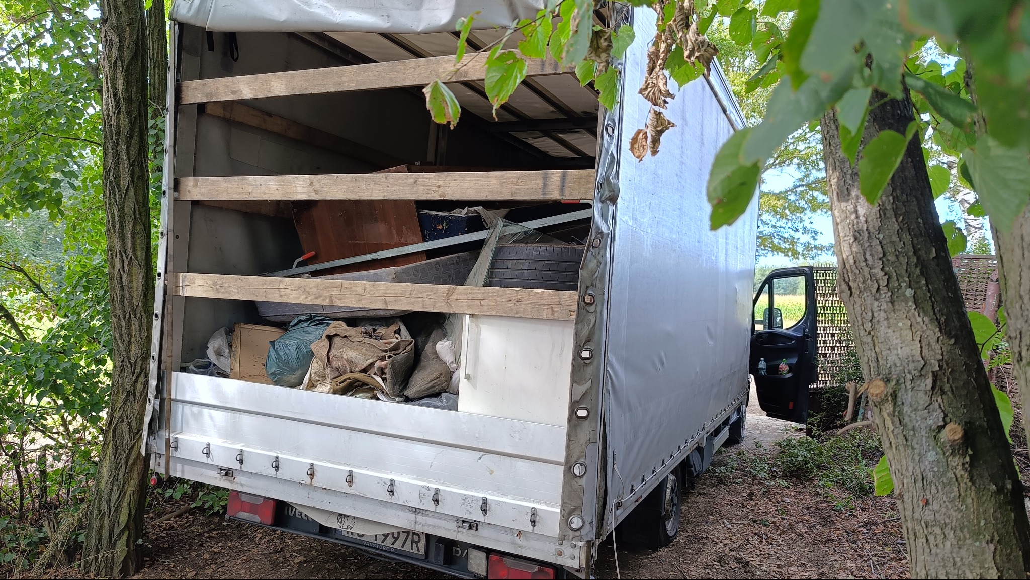 Otwarta paka ciężarówki wypełniona różnymi przedmiotami, widoczna przez drzewa. Częściowo załadowany pojazd sugeruje etap załadunku podczas przeprowadzki.