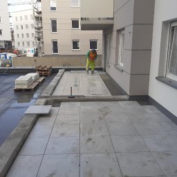 EX-BUD 78 SPÓŁKA Z OGRANICZONĄ ODPOWIEDZIALNOŚCIĄ - Układanie szarych płyt chodnikowych na tarasie budynku wielorodzinnego. Prace wykończeniowe z widocznymi elementami konstrukcyjnymi i pracownikami w odblaskowych ubraniach.
