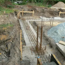 Zbrojenie fundamentów w wykopie, przygotowane do zalania betonem. Widoczny żwir, drewniane szalunki i wykopana ziemia. Prace budowlane w toku.