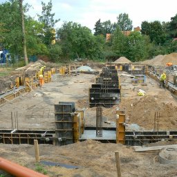 EX-BUD 78 SPÓŁKA Z OGRANICZONĄ ODPOWIEDZIALNOŚCIĄ - Wykopane fundamenty domu z częściowo zalanym betonem i szalunkami. Widoczni robotnicy w żółtych kaskach. Na pierwszym planie rura kanalizacyjna.