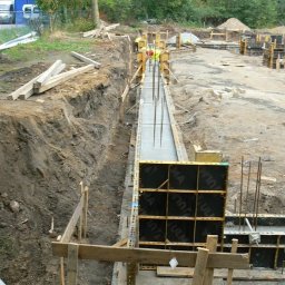 EX-BUD 78 SPÓŁKA Z OGRANICZONĄ ODPOWIEDZIALNOŚCIĄ - Wykop z deskowaniem i zalewaniem betonem, widoczne zbrojenie pionowe. W tle drzewa i zabudowa mieszkalna. Prace ziemne w toku, etap budowy fundamentów.