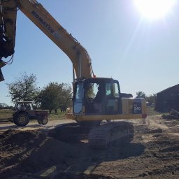 EX-BUD 78 SPÓŁKA Z OGRANICZONĄ ODPOWIEDZIALNOŚCIĄ - Żółta koparka Komatsu PC 240 LC w trakcie prac ziemnych na placu budowy. W tle widoczny traktor i budynek gospodarczy. Słoneczny dzień.