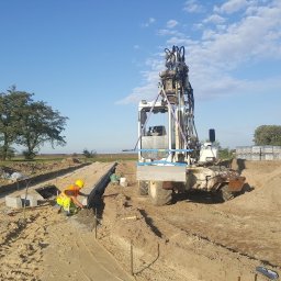 EX-BUD 78 SPÓŁKA Z OGRANICZONĄ ODPOWIEDZIALNOŚCIĄ - Układanie obrzeży betonowych przez pracownika w żółtym kasku, z użyciem poziomicy na wykopie, w tle maszyna budowlana i zielone pole.