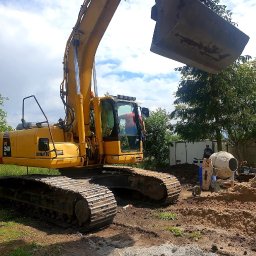 EX-BUD 78 SPÓŁKA Z OGRANICZONĄ ODPOWIEDZIALNOŚCIĄ - Żółta koparka Komatsu PC 240 LC z podniesioną łyżką na tle zieleni, obok betoniarka i pracownik. Wykop ziemi na budowie w słoneczny dzień.