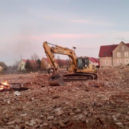 EX-BUD 78 SPÓŁKA Z OGRANICZONĄ ODPOWIEDZIALNOŚCIĄ - Żółta koparka Komatsu na gruzowisku po rozbiórce budynku, w tle zabudowania wiejskie i palące się ognisko, pochmurne niebo.