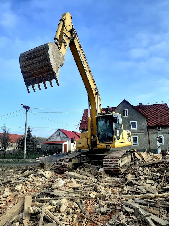 Żółta koparka Komatsu na gruzowisku po rozbiórce budynku. W tle budynki mieszkalne i słup z gniazdem bocianim. Błękitne niebo.