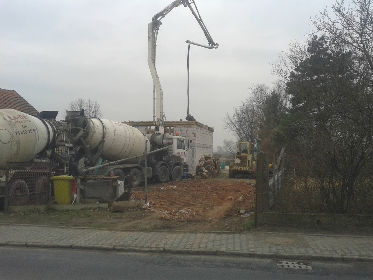Wylewanie betonu na budowę domu jednorodzinnego. Dwa betonomieszarki i pompa do betonu w akcji, w tle widoczny budynek w stanie surowym otwartym.