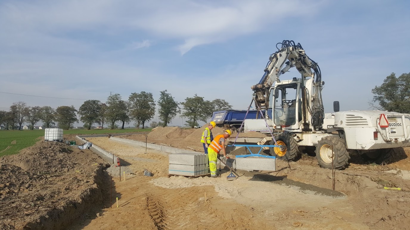Układanie kostki brukowej na przygotowanym podłożu przez robotników w żółtych kamizelkach. W tle maszyna budowlana i wywrotka. Praca w toku.