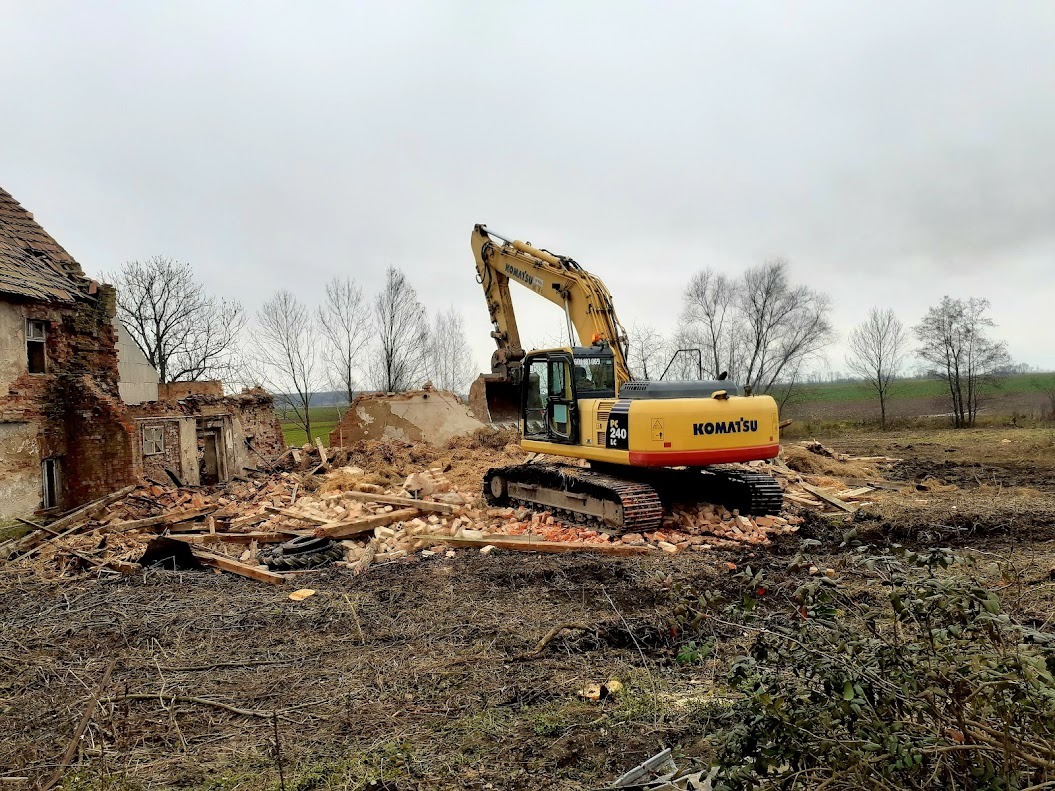Żółta koparka Komatsu PK 240 na gruzowisku po rozbiórce ceglanego budynku, z fragmentem ściany i drzewami w tle, na tle pochmurnego nieba.