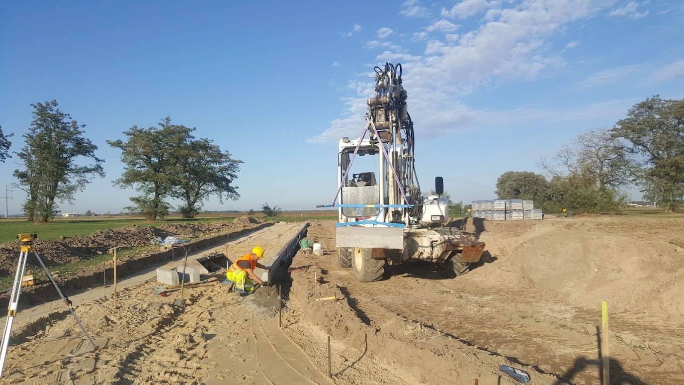 Układanie obrzeży betonowych w wykopie pod drogę, pracownik w kamizelce, poziomica, maszyna do układania obrzeży. Etap budowy drogi na tle zieleni i błękitnego nieba.