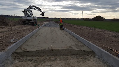 Utwardzanie terenu pod drogę: pracownik z zagęszczarką, obramowanie z betonowych krawężników, w tle koparka i pole uprawne pod pochmurnym niebem.