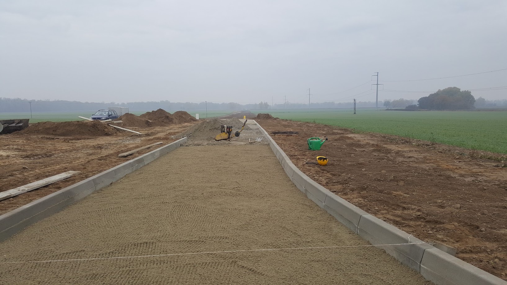 Budowa drogi gruntowej z ułożonymi krawężnikami. W tle widać sprzęt budowlany, hałdy ziemi i pola uprawne pod pochmurnym niebem.
