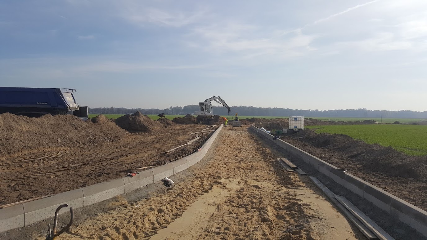 Budowa drogi gruntowej z ułożonymi obrzeżami betonowymi. W tle koparka i wywrotka, widoczne ślady opon na piasku. Prace drogowe w toku, słoneczny dzień.