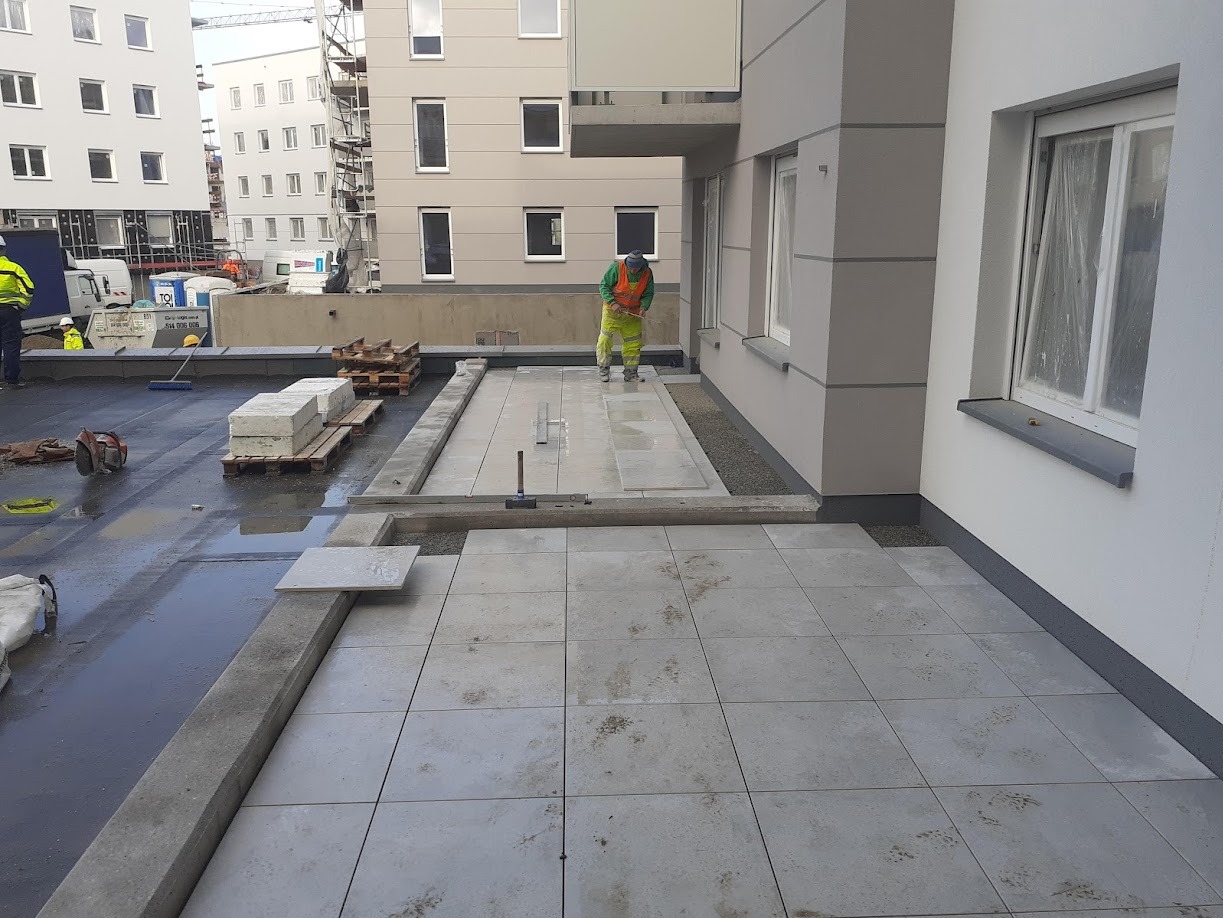 Układanie szarych płyt chodnikowych na tarasie budynku wielorodzinnego. Prace wykończeniowe z widocznymi elementami konstrukcyjnymi i pracownikami w odblaskowych ubraniach.