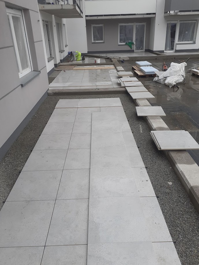 Układanie jasnoszarej kostki brukowej na zewnątrz budynku. Widoczny pracownik w odblaskowej kamizelce, częściowo ułożona nawierzchnia i przygotowane płyty.