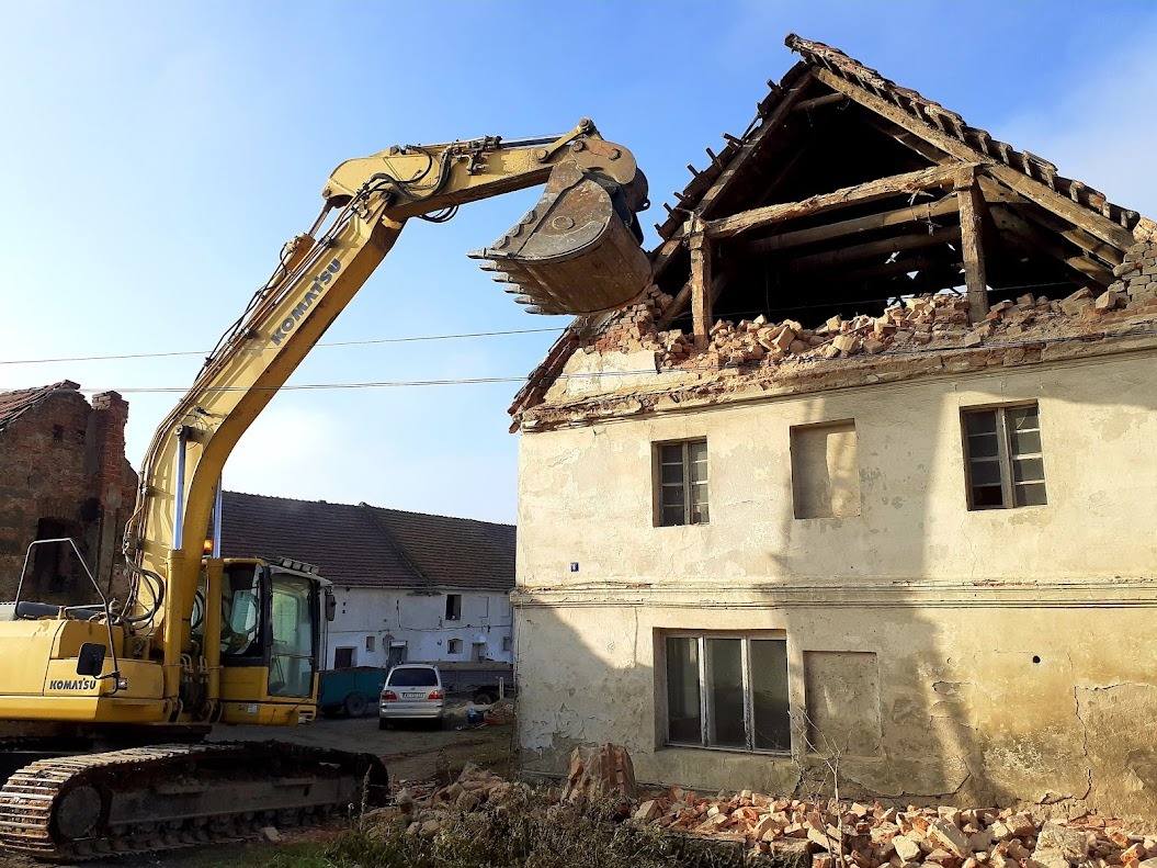 Żółta koparka Komatsu podczas rozbiórki starego, dwupiętrowego budynku. Częściowo zburzony dach z widocznymi belkami i gruzem. W tle budynki gospodarcze i niebo.