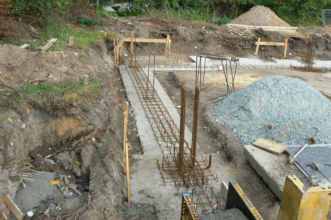 Zbrojenie fundamentów w wykopie, przygotowane do zalania betonem. Widoczny żwir, drewniane szalunki i wykopana ziemia. Prace budowlane w toku.