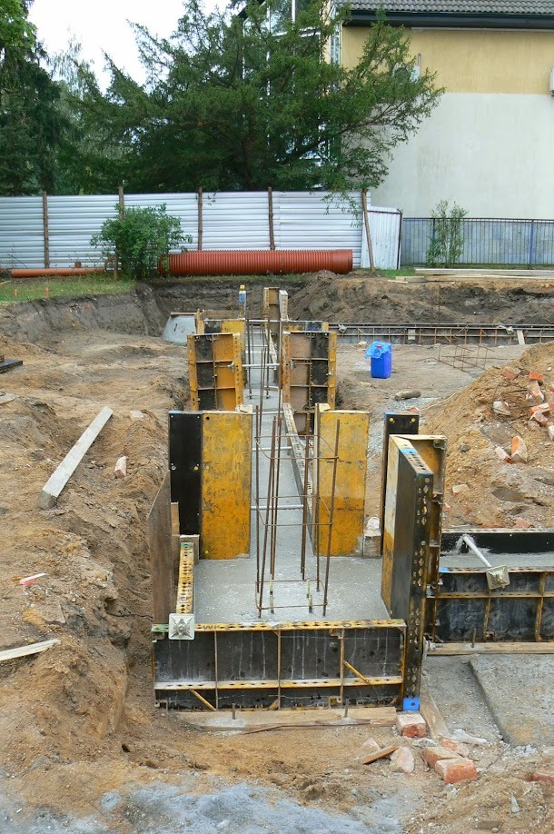 Zalewanie fundamentów zbrojonych w szalunkach. Widok na plac budowy w Warszawie. W tle widoczny płot z blachy falistej i fragment budynku.