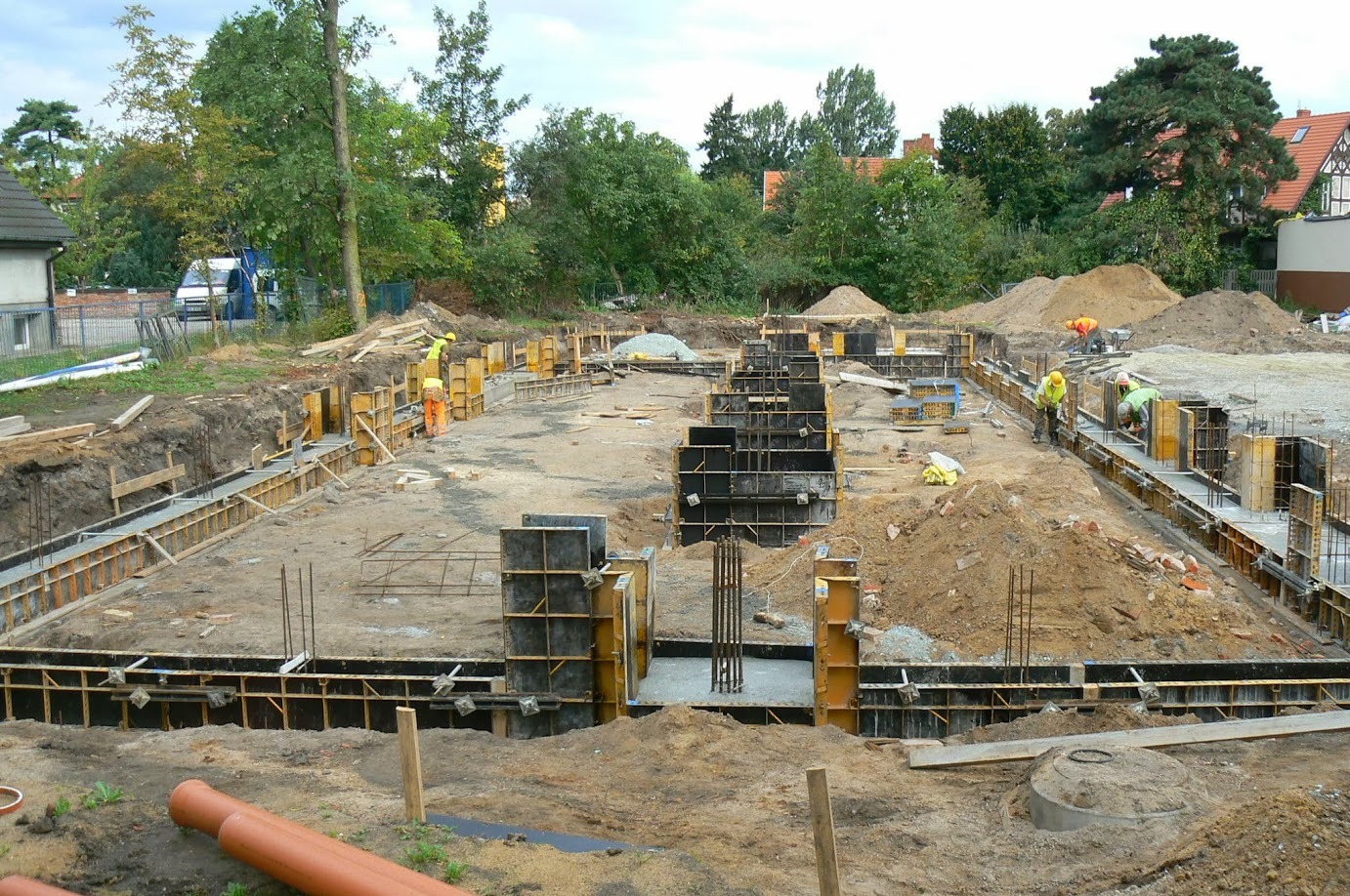 Wykopane fundamenty domu z częściowo zalanym betonem i szalunkami. Widoczni robotnicy w żółtych kaskach. Na pierwszym planie rura kanalizacyjna.