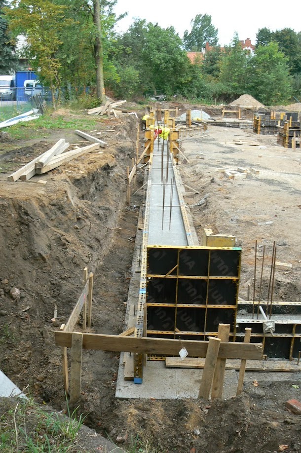 Wykop z deskowaniem i zalewaniem betonem, widoczne zbrojenie pionowe. W tle drzewa i zabudowa mieszkalna. Prace ziemne w toku, etap budowy fundamentów.