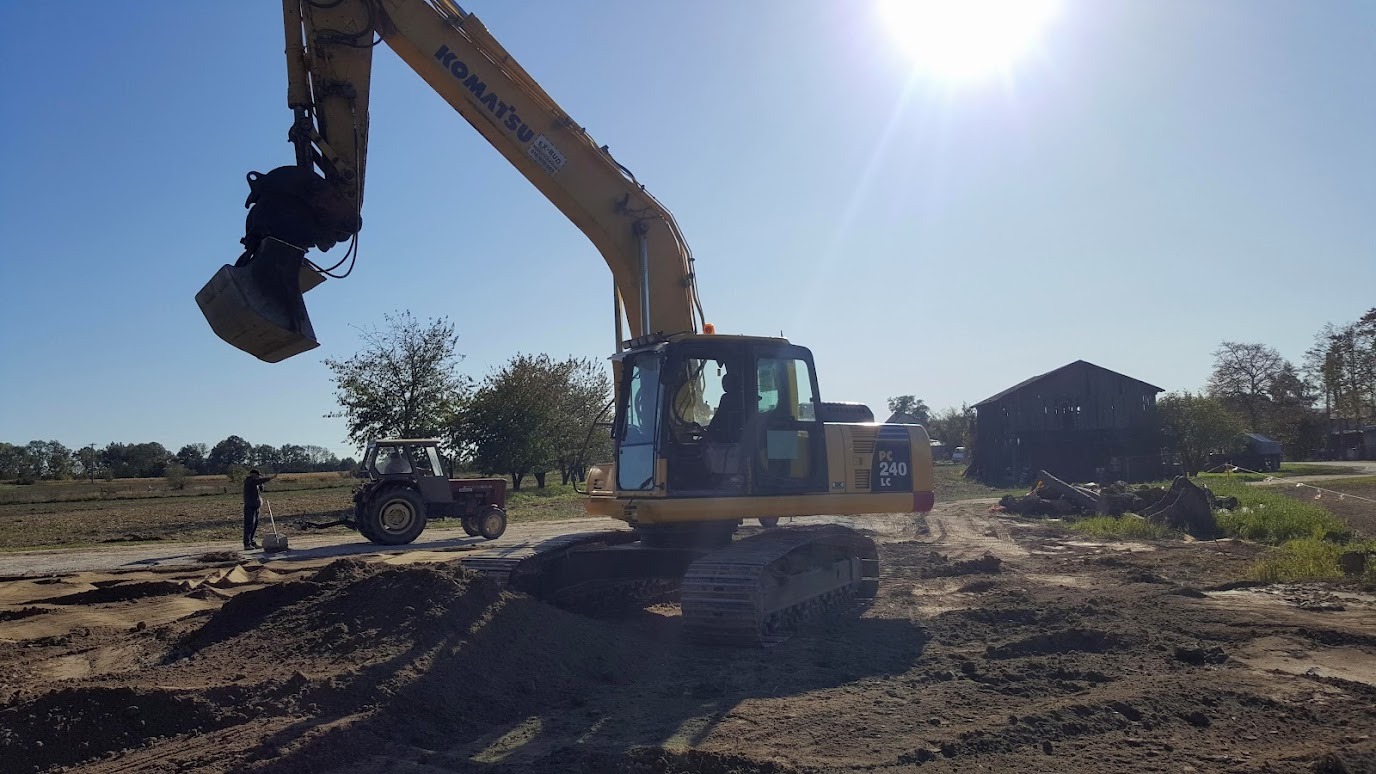 Żółta koparka Komatsu PC 240 LC w trakcie prac ziemnych na placu budowy. W tle widoczny traktor i budynek gospodarczy. Słoneczny dzień.