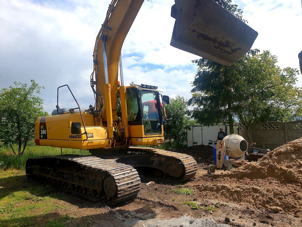 Żółta koparka Komatsu PC 240 LC z podniesioną łyżką na tle zieleni, obok betoniarka i pracownik. Wykop ziemi na budowie w słoneczny dzień.