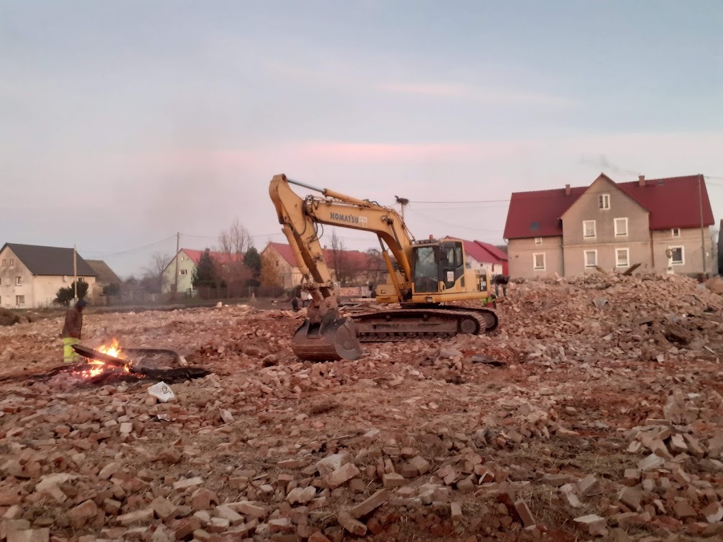 Żółta koparka Komatsu na gruzowisku po rozbiórce budynku, w tle zabudowania wiejskie i palące się ognisko, pochmurne niebo.