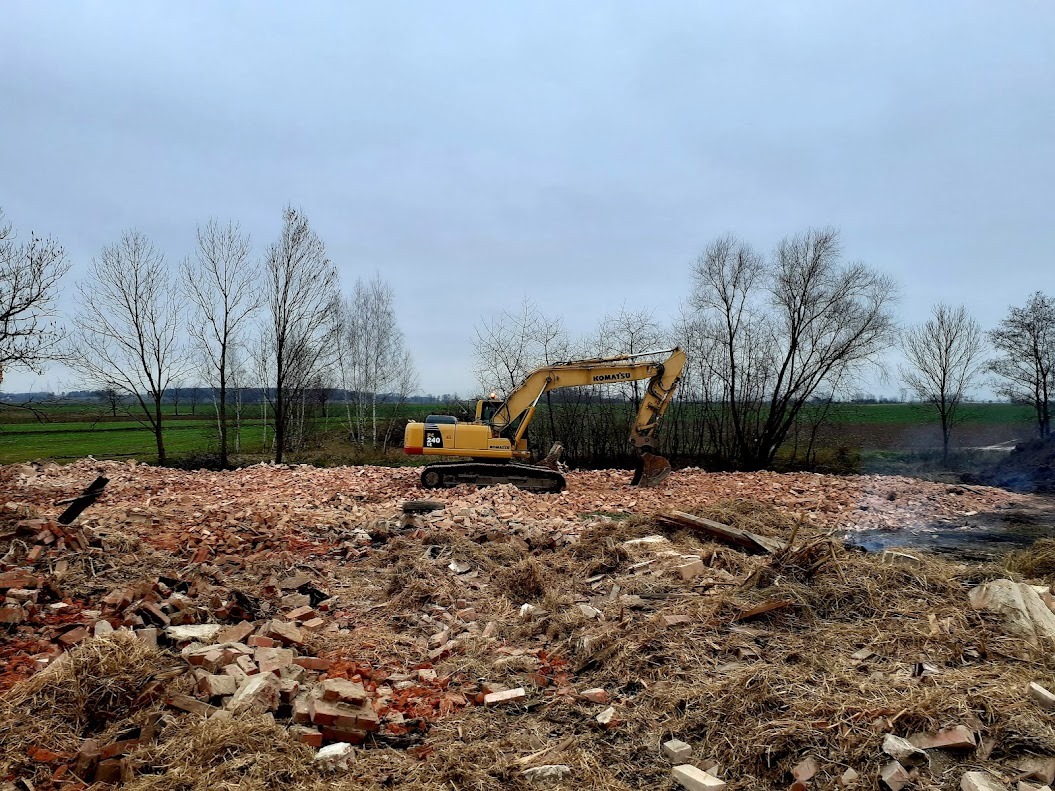 Żółta koparka Komatsu na tle gruzu z cegieł i resztek roślinnych, po rozbiórce budynku. W tle widoczne drzewa i pole pod pochmurnym niebem.