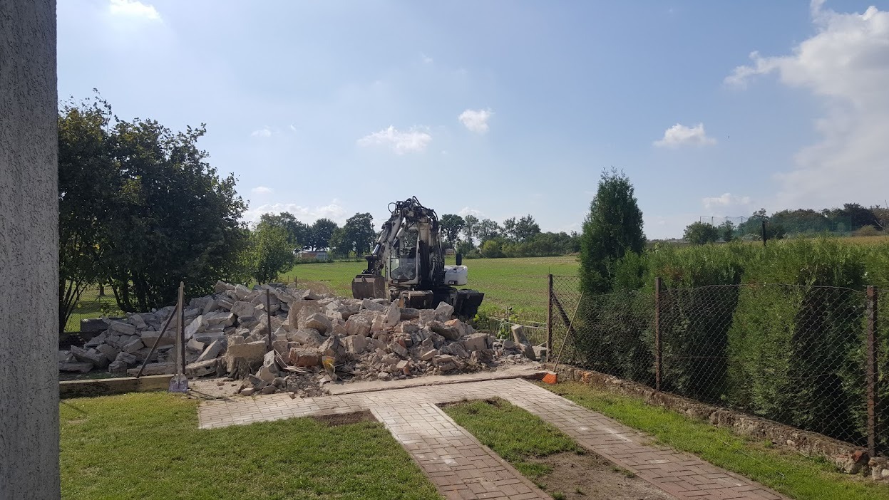 Koparka na działce z gruzem po rozbiórce, widok z boku domu. Na pierwszym planie trawnik i ścieżka z kostki, w tle drzewa i pole pod błękitnym niebem.