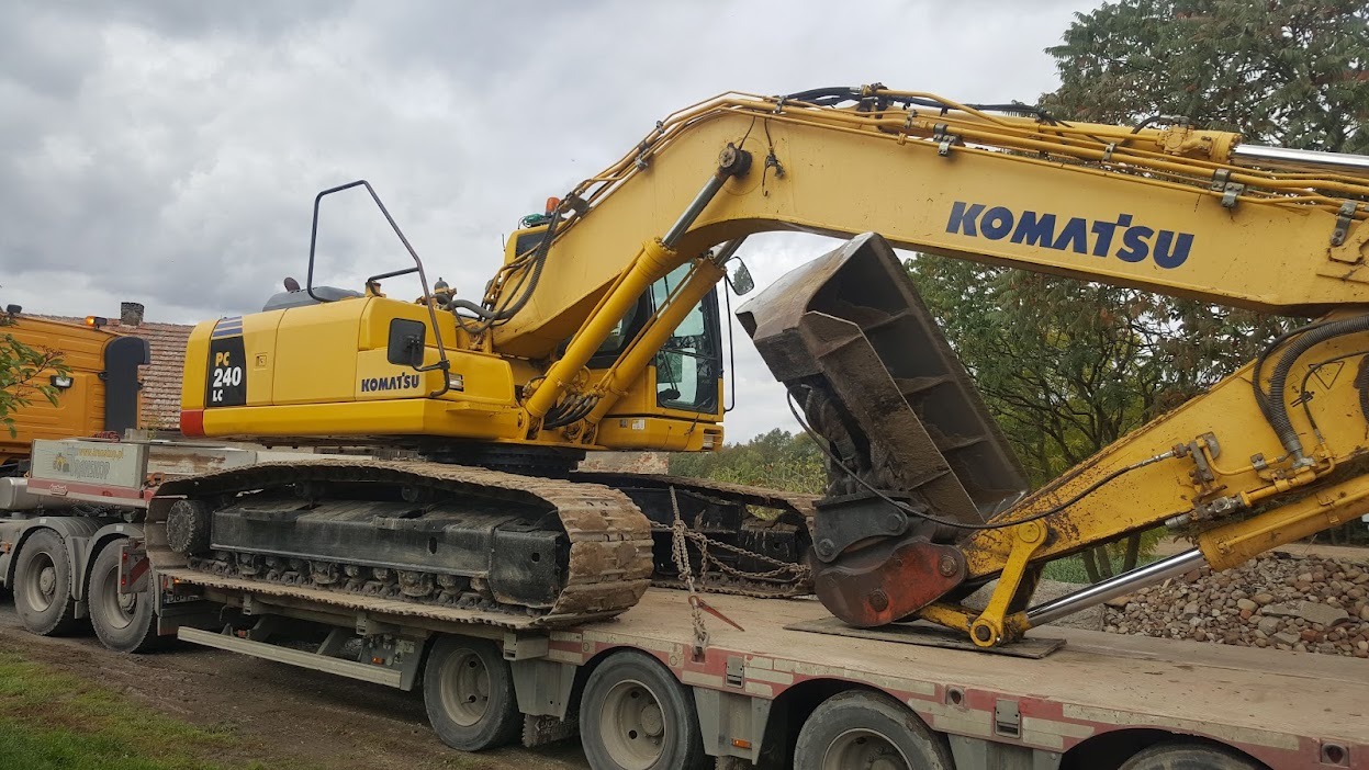 Żółta koparka Komatsu PC240LC na niskopodwoziowej przyczepie. Widoczne gąsienice, ramię z łyżką i fragment kabiny. W tle drzewa i budynek.
